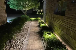 Well-lit stone pathway alongside a brick house, accented by soft landscape lighting and green shrubs at night.