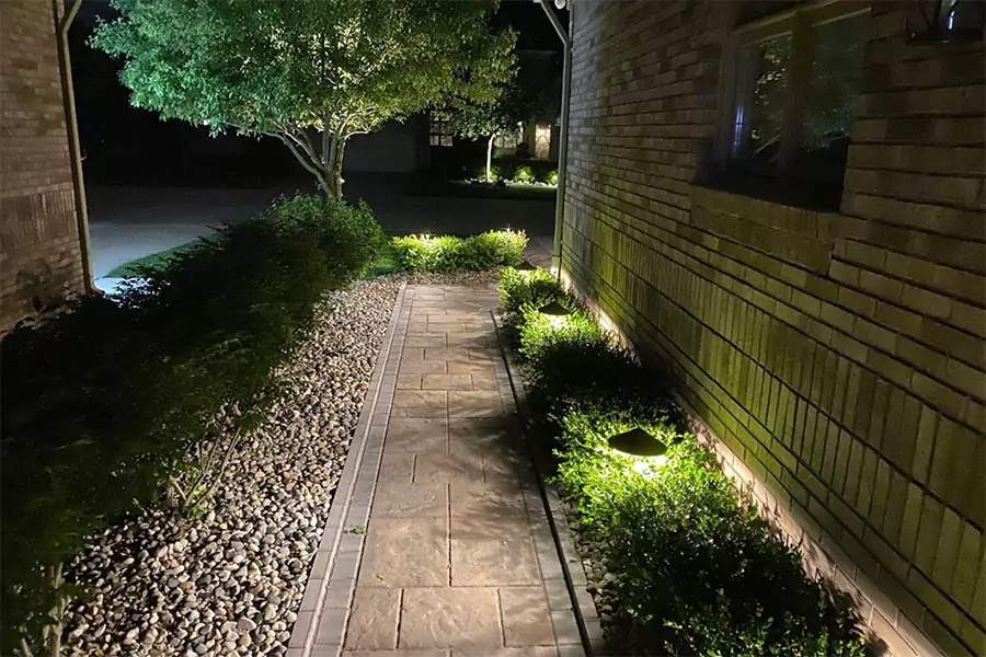 Well-lit stone pathway alongside a brick house, accented by soft landscape lighting and green shrubs at night.