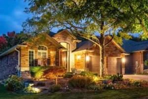 Luxurious home at night with driveway and landscape illuminated by warm, strategic outdoor lighting, highlighting trees, flower beds, and the house facade.