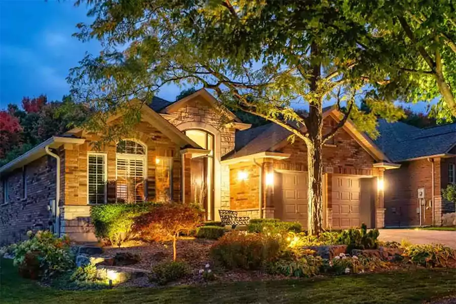 Luxurious home at night with driveway and landscape illuminated by warm, strategic outdoor lighting, highlighting trees, flower beds, and the house facade.