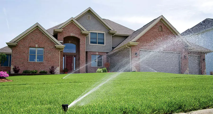 Lawn sprinkler system watering a lush green yard in front of a brick home—illustrating efficient irrigation system use for summer lawn care in Humble, TX.